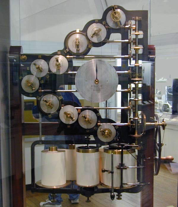 A tall wooden frame behind glass holding about a dozen white-faced brass wheels on levers, with a large dial at the center and three paper rolls at the bottom