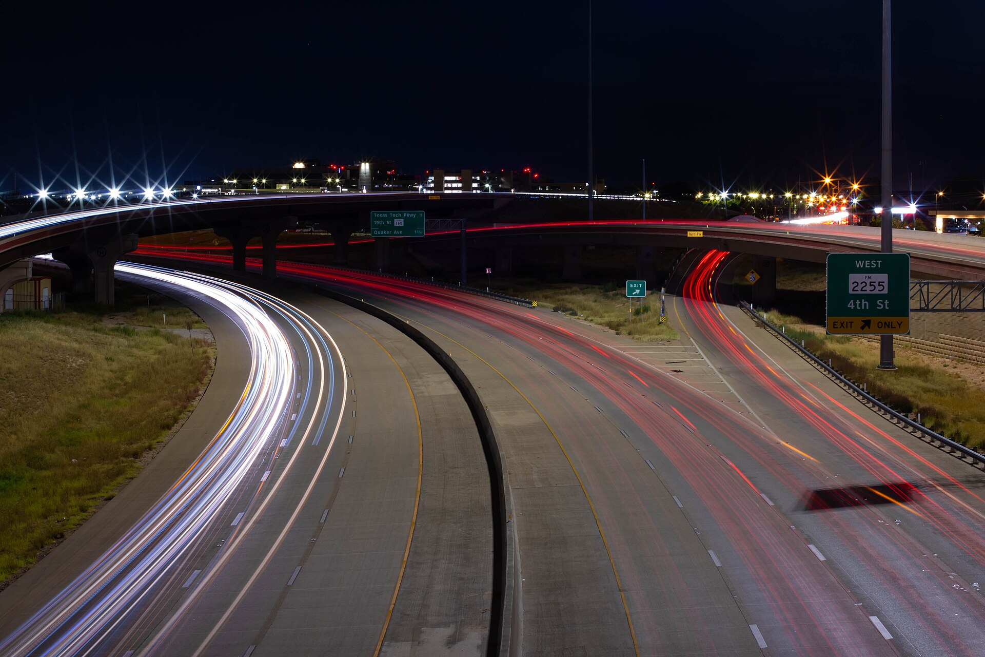 A curving Texas freeway at night, shot from an overpass with the shutter held open. Headlights stretch into white ribbons on the near lanes and taillights into red ribbons on the far lanes, under an exit sign for 4th Street