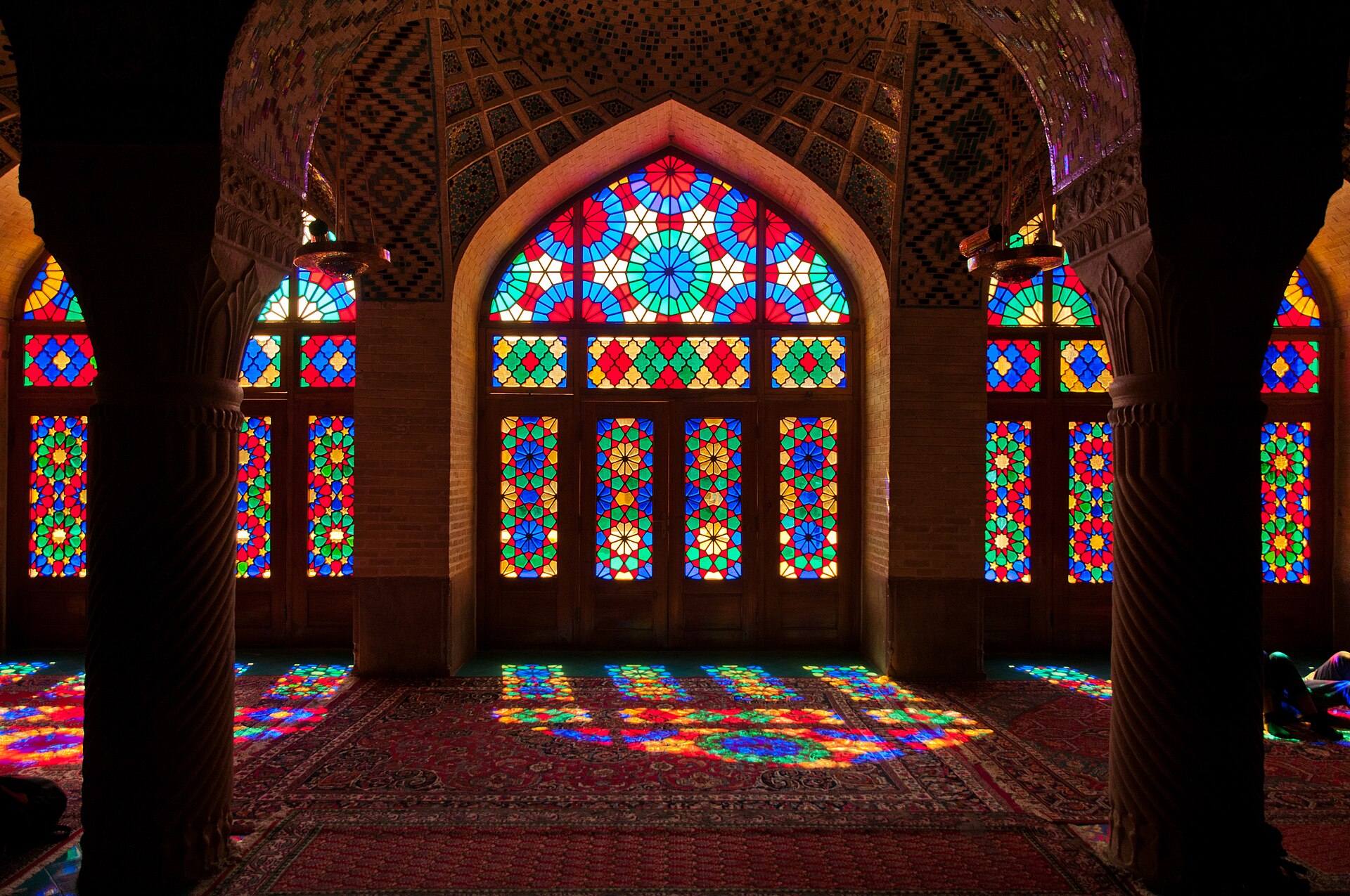 An arched wall of red, blue, yellow and green glass set in a wooden lattice, inside a prayer hall with dark columns. Sunlight through the glass lays the same lattice, in color, across the carpet