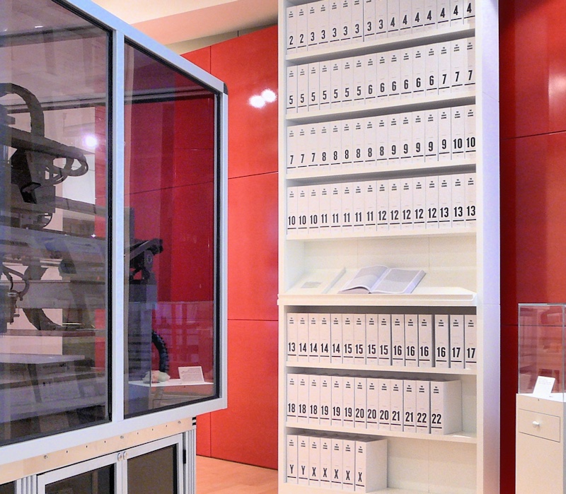 A tall white bookcase against a red wall, its shelves packed with matching white volumes. Each spine carries a large black number from 1 to 22, then X and Y, and one book lies open on a middle shelf