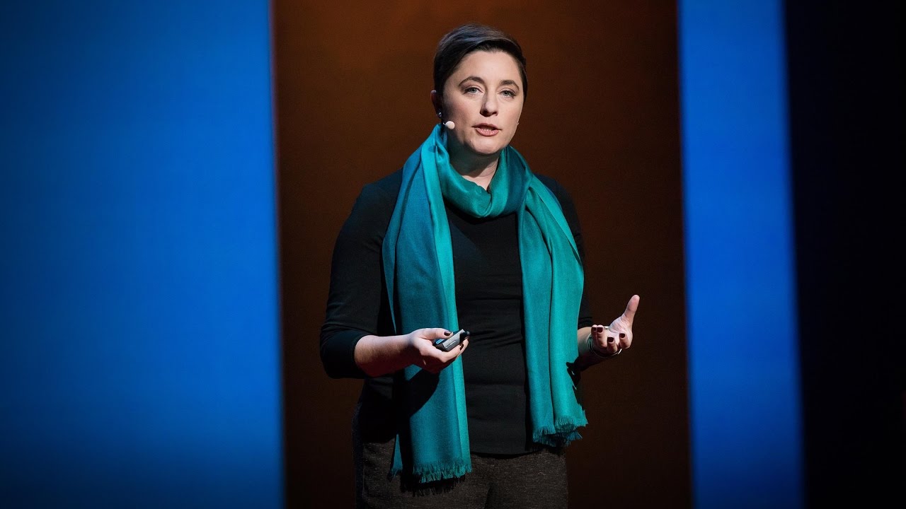 A woman with short dark hair and a teal scarf speaks on a dark stage between two tall blue panels, a slide clicker in one hand