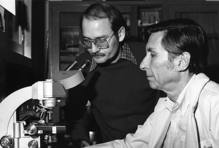 Black-and-white photo of two men at a laboratory microscope, the younger in glasses looking into the eyepieces, the older in a white lab coat beside him