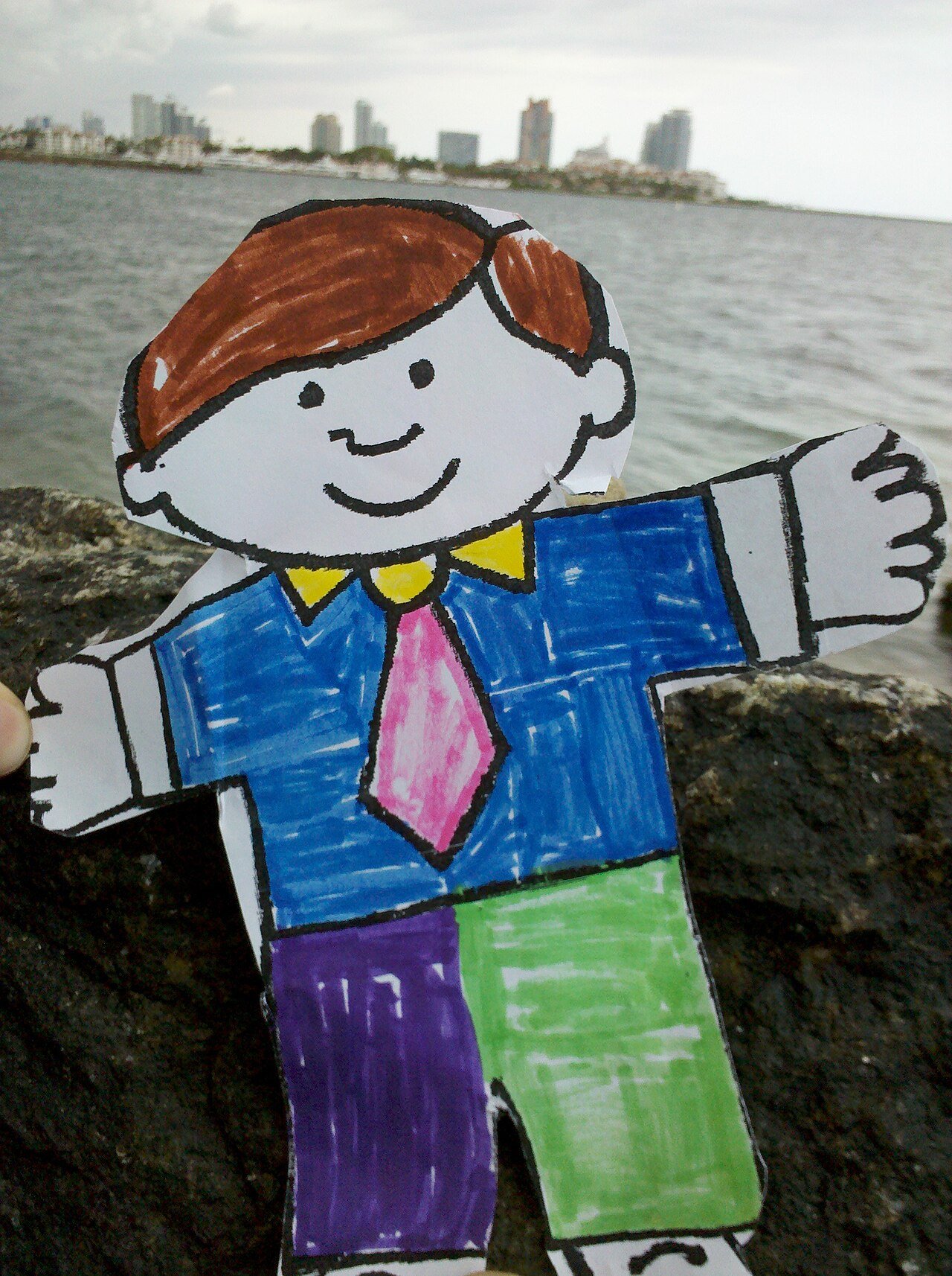 A hand-colored paper cutout of Flat Stanley, in a blue shirt, pink tie and green trousers, propped on a rock with the Miami Beach skyline across the water behind him