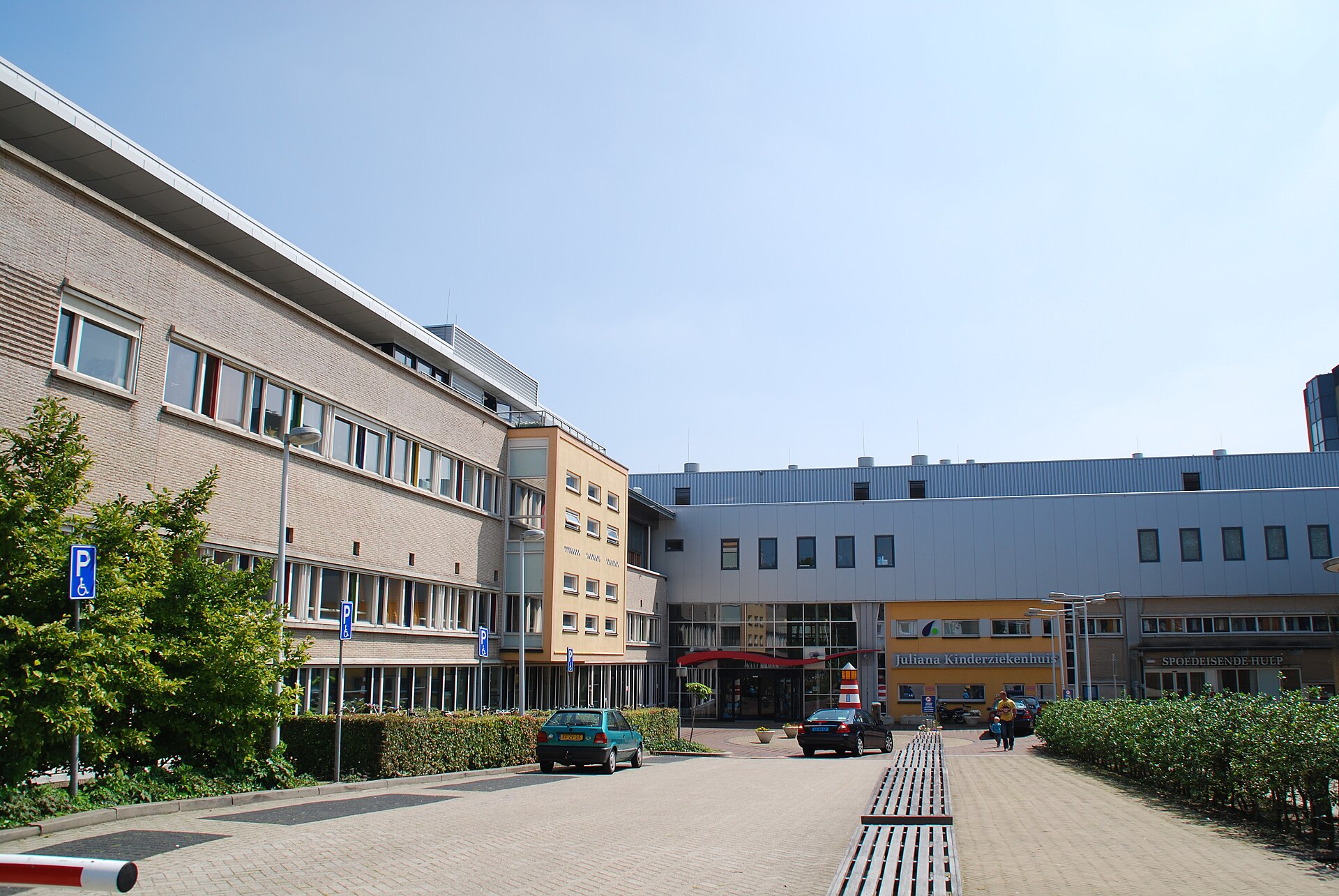 A long brick and grey hospital building in sunshine, a paved forecourt with benches and two parked cars in front